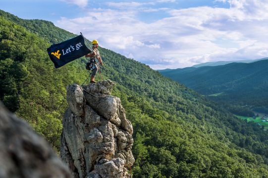 A student on top of mountain holding a WVU branded flag.