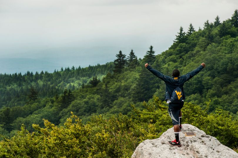 A student on a cliff raising is hands excitingly with a WVU branded backpack on.